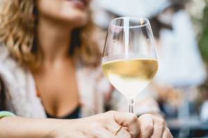 Glas Weißwein in der Hand, im Hintergrund Frau bei Weinprobe im Freien