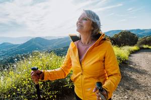 Lächelnde Seniorin wandert mit Trekkingstöcken durch blühende Berglandschaft