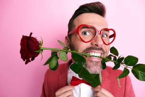 Lächelnder Mann mit Herzbrille hält rote Rose im Mund, Valentinstag-Motiv