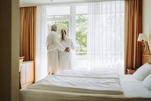 Paar in Bademänteln blickt aus Hotelzimmerfenster ins Grüne, Thermenhotel Sonnenhof